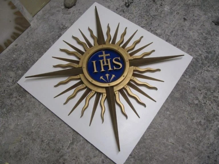 A decorative sunburst wall piece with gold rays and a blue circle in the center that has the letters 'IHS' in gold.  Creative and Custom fibreglass and composite solutions. 