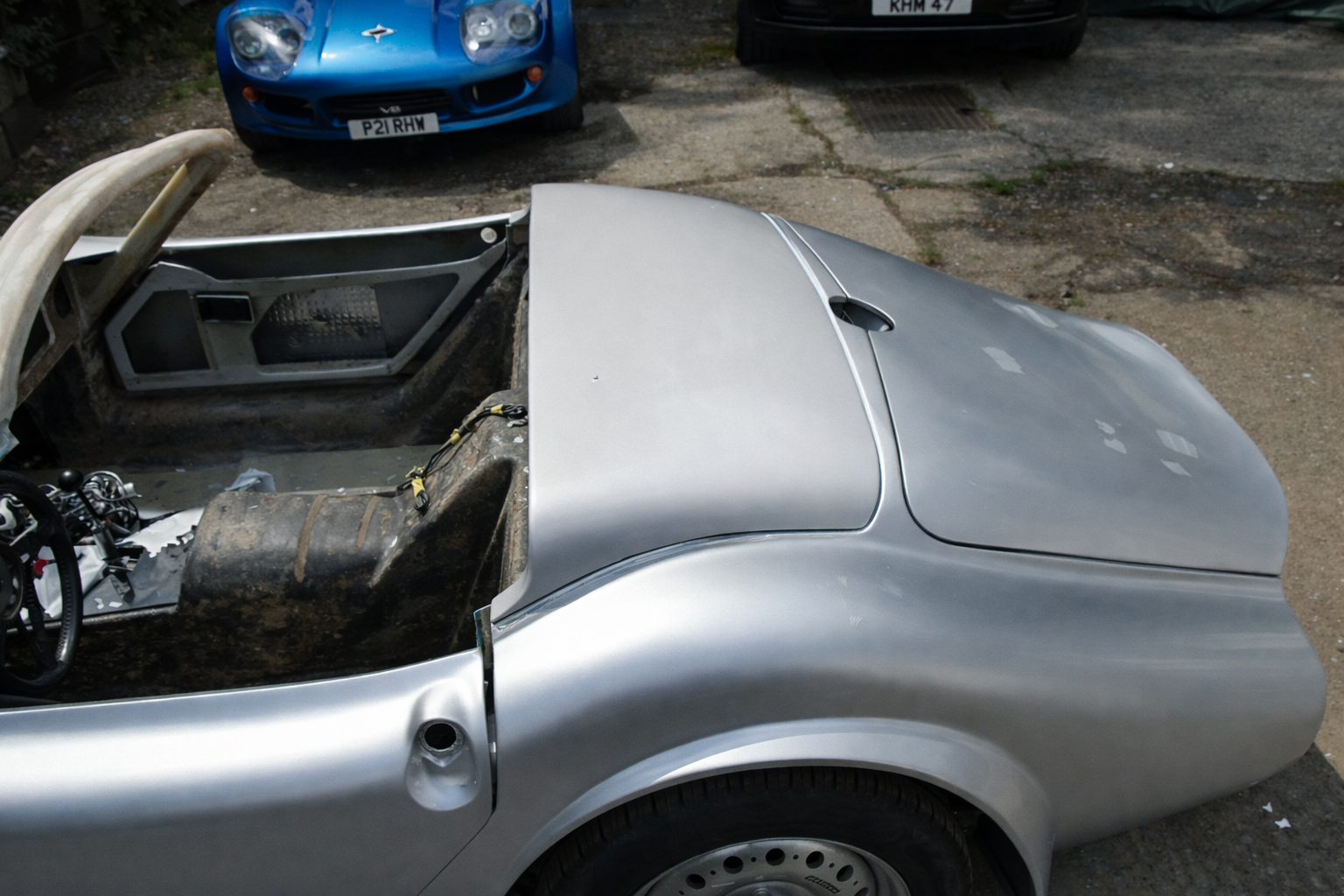 Close-up of a silver sports car without a windshield, showing exposed interior and missing parts, parked on a dirty ground. A blue car is visible in the background.  Technical Composites.