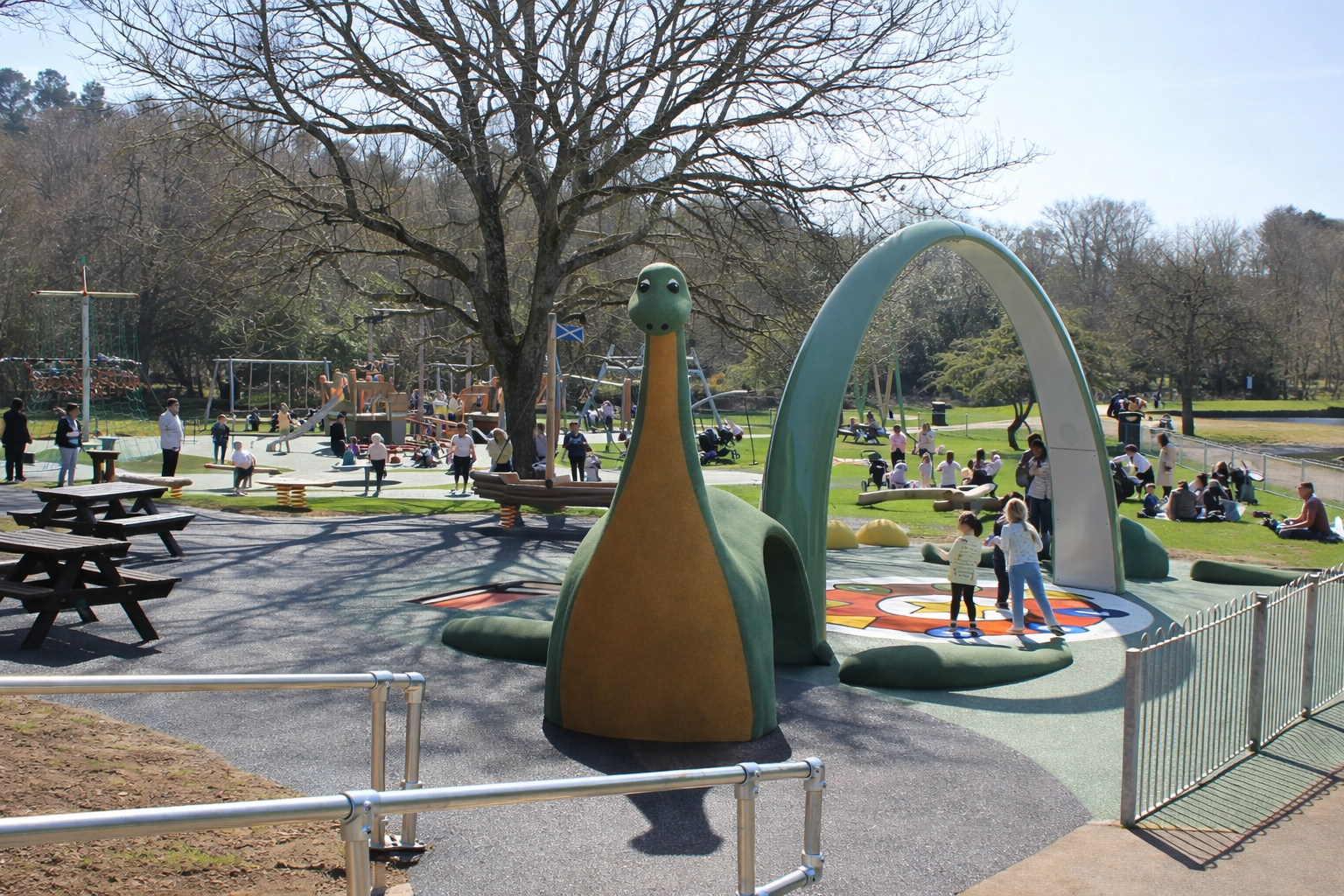 Children playing on a playground shaped like the Loch Ness Monster in a park with trees and many people sitting and walking on the grass. Creative and Custom fibreglass and composite solutions.