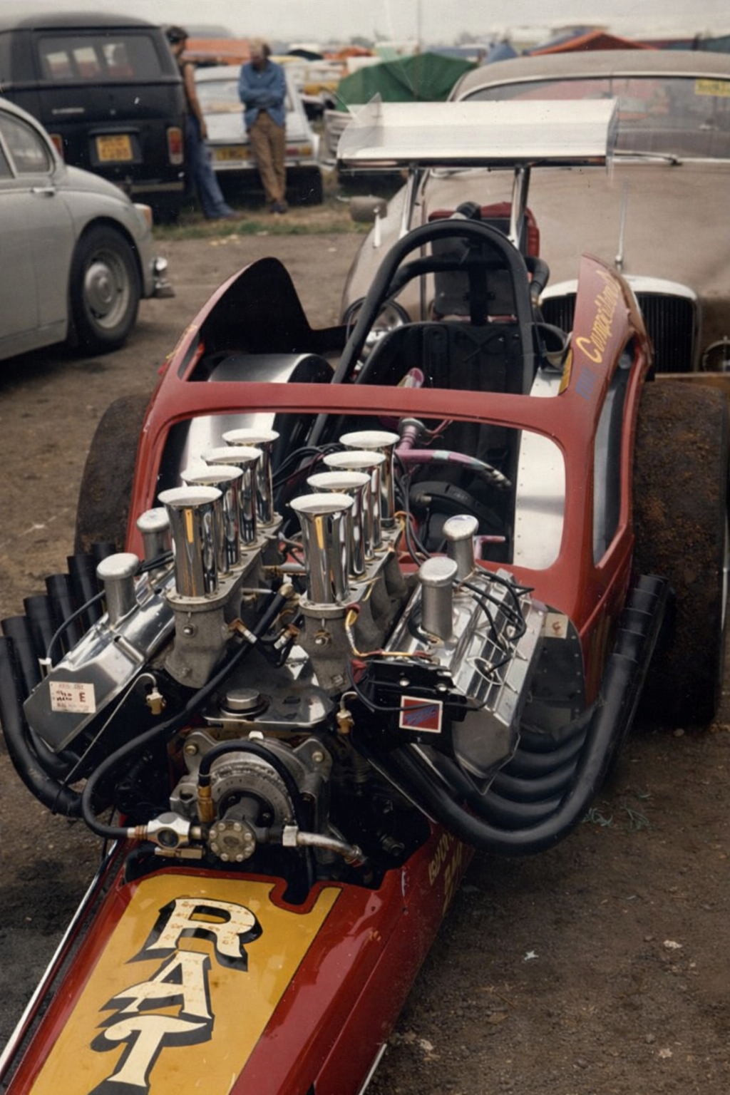 Close-up of a vintage race car engine with chrome exhaust pipes and a red and yellow body, labeled 'RAT', in a dirt lot with vintage cars and people in the background.