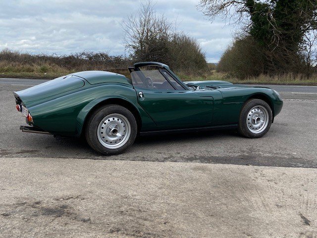 A classic dark green roadster with a convertible top parked on the side of a road against a background of trees and cloudy sky.  Technical Composites.