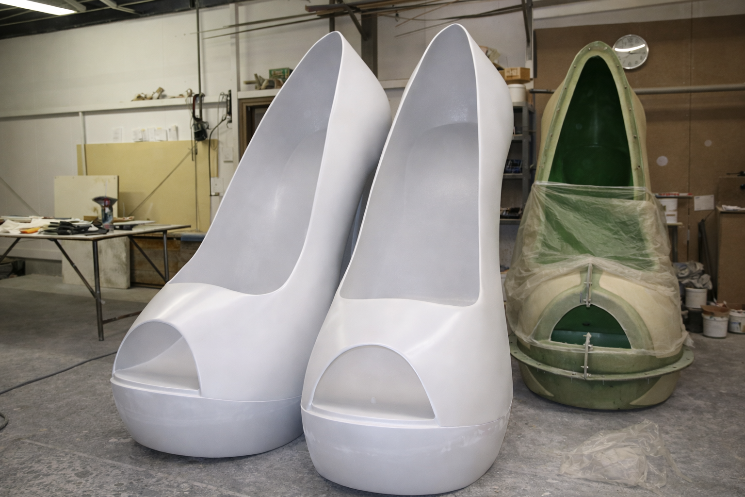 Two large white high-heeled shoe sculptures with open toes in a workshop setting, with an unfinished boat hull covered with plastic behind them. Creative and Custom fibreglass and composite solutions.