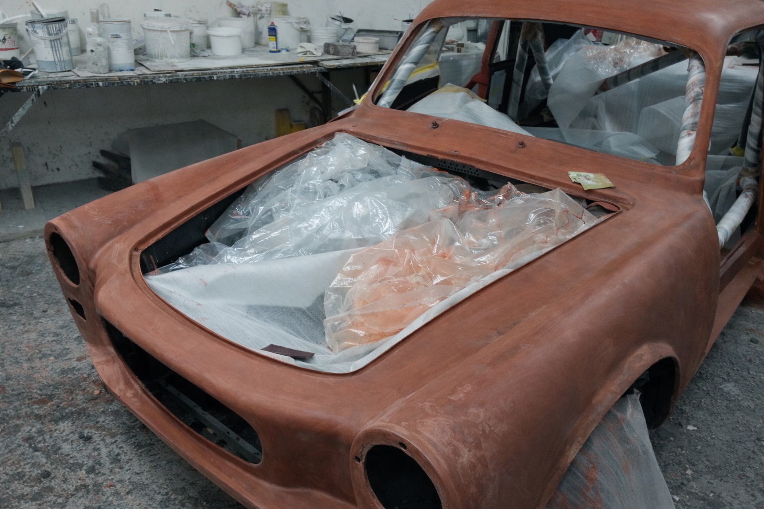 A vintage car body undergoing restoration with a rusty, unfinished surface, covered with protective plastic sheeting inside a workshop filled with tools and paint supplies.  Technical Composites.