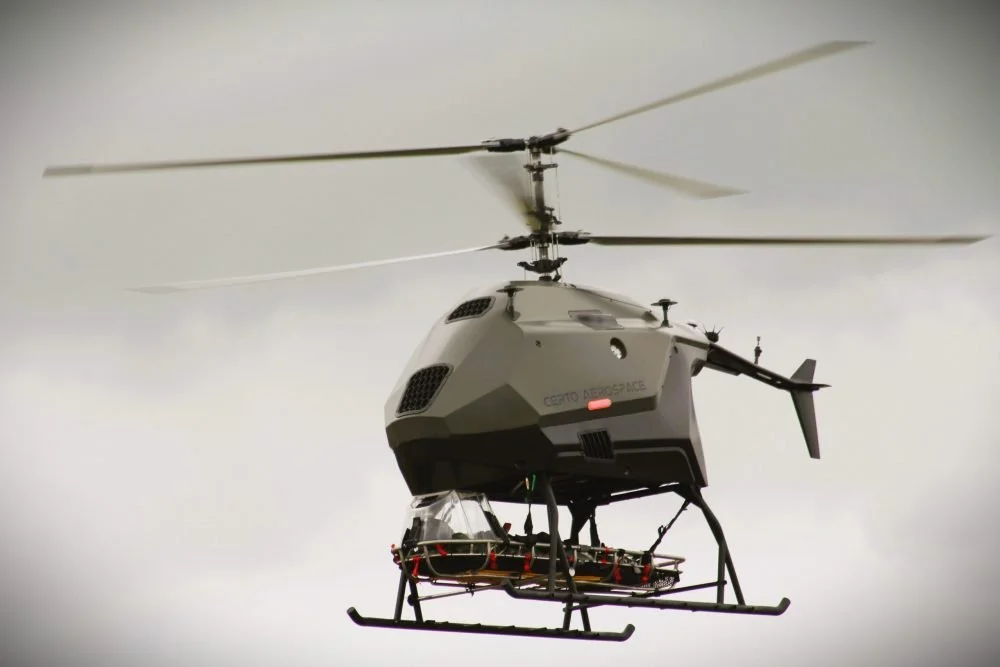 CERTO A military-style drone helicopter with a mounted camera or sensor system underneath, flying in a cloudy sky.  Technical Composites for aerospace.