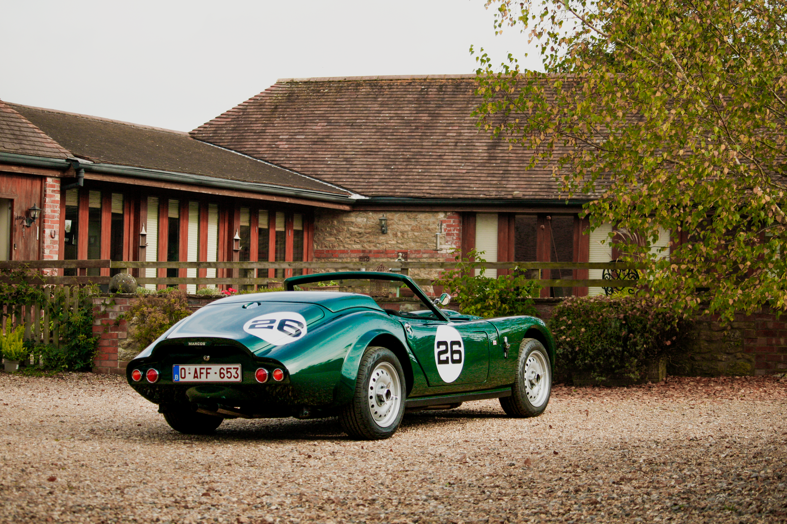 A vintage green race car with the number 26 on the door and trunk, parked on a gravel driveway in front of a house with a brick and wood exterior, surrounded by greenery.  Technical Composites.