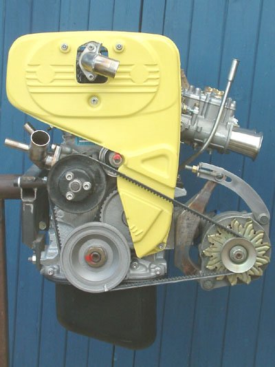 A small gray engine with a yellow cover and a belt-driven pulley system mounted on a blue wooden wall.  Technical Composites.
