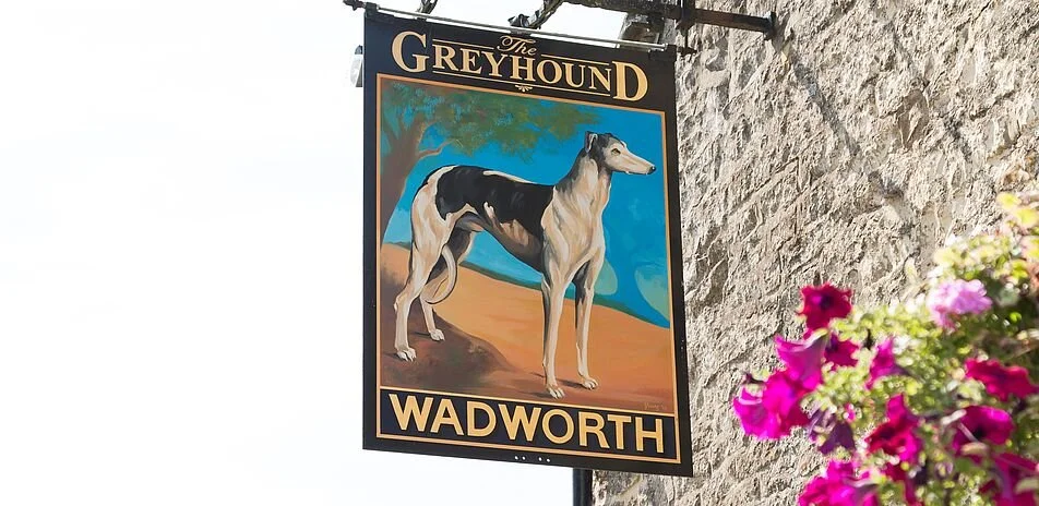 Wadsworth pub sign for The Greyhound, a pub or establishment, featuring a painting of a greyhound dog, mounted on a stone wall with pink and purple flowers nearby.  Creative and Custom fibreglass and composite solutions. 