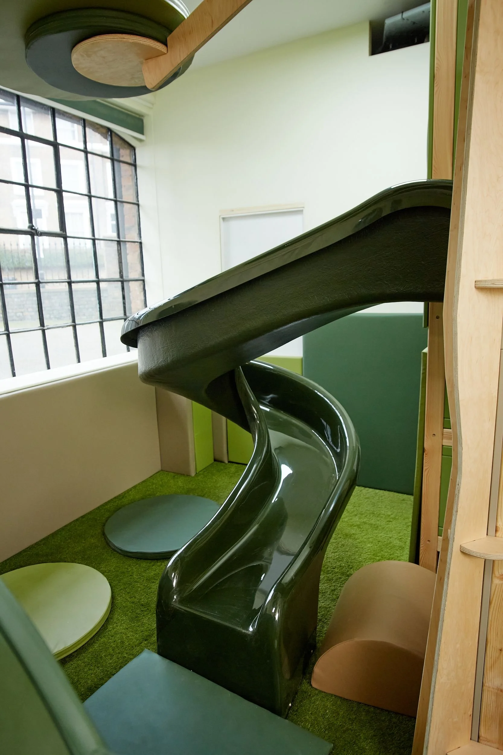 Indoor playground slide made of fibreglass, set on green artificial grass, with adjacent padded seats in pastel colours, in a room with large windows and light green walls. Creative and Custom fibreglass and composite solutions.