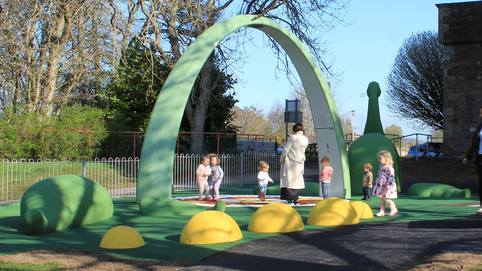 Children playing at a modern, whimsical playground with a large green arch and Loch Ness Monster fibreglass structures on a sunny day. Creative and Custom fibreglass and composite solutions.