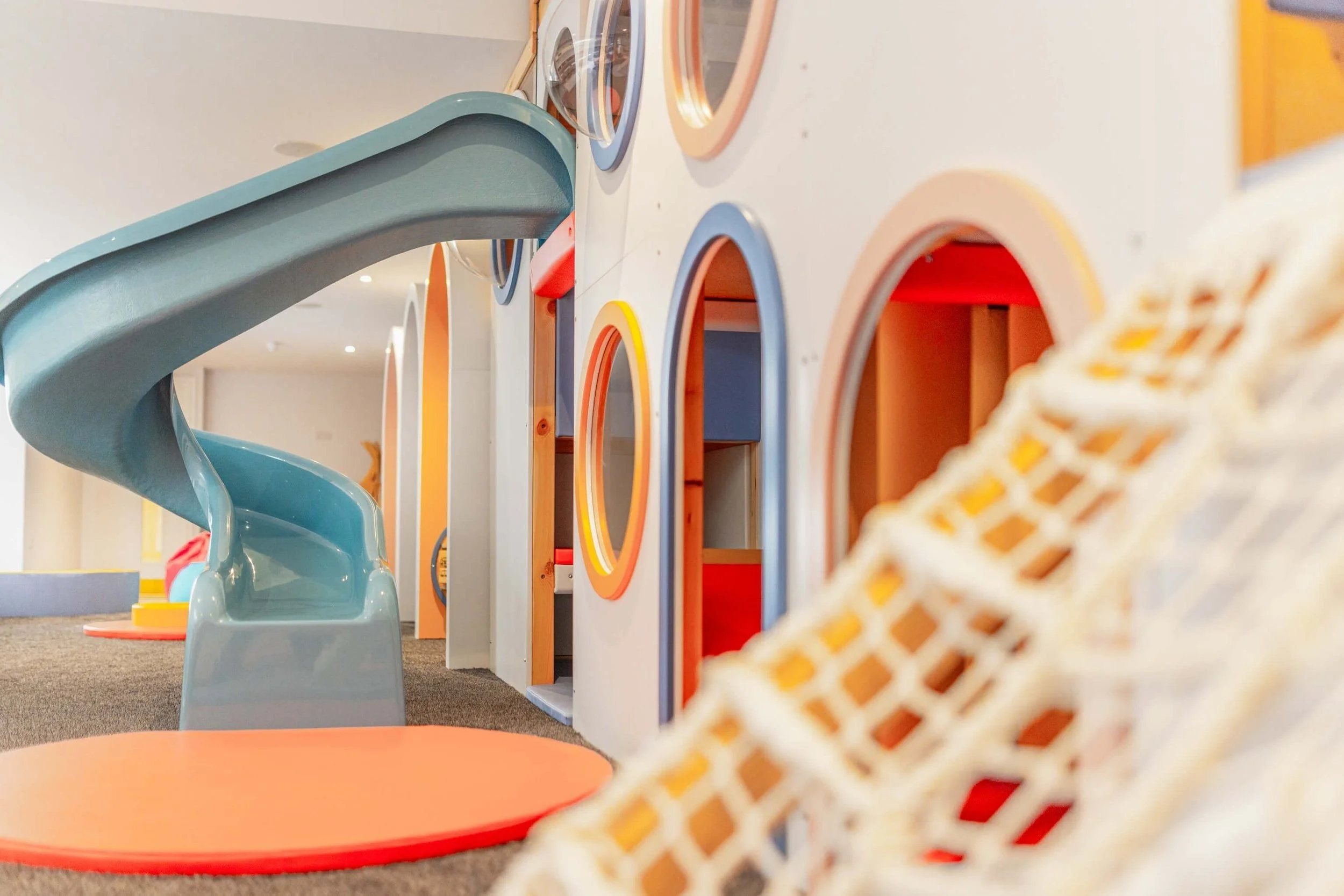 Colorful indoor playground with a blue spiral slide, geometric structures, and padded flooring.