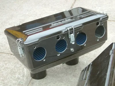 Black automotive engine valve cover with multiple circular openings and blue vent filters, placed on a concrete surface.  Technical Composites.