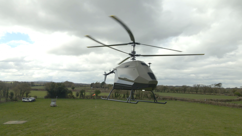 CERTO A white and gray helicopter flying low over a grassy field with a windsock and a small vehicle in the background under a cloudy sky.  Technical Composites for aerospace.