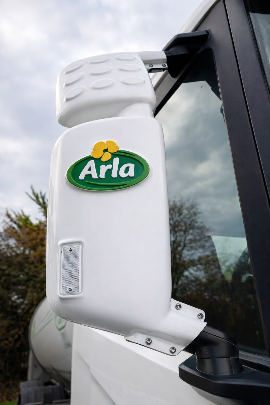 A close-up of an Arla-branded milk tanker truck mirror with the Arla logo and a yellow flower, seen outdoors against a cloudy sky.