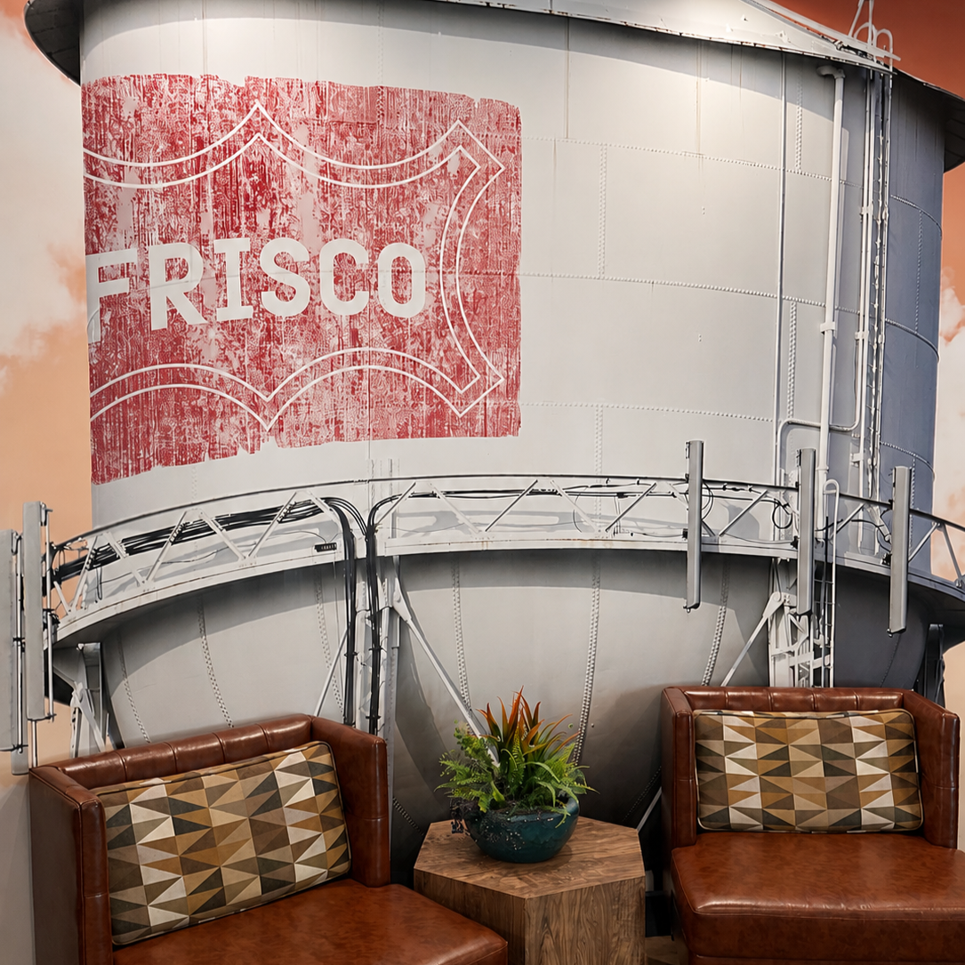 Interior of a room with a large industrial water tank in the background, featuring a faded red and white "Fresco" sign, and two brown leather chairs flanking a small wooden table with a green potted plant.