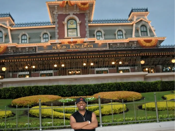 Person standing in front of Disneyland's Magic Kingdom entrance with a garden and fence in foreground.