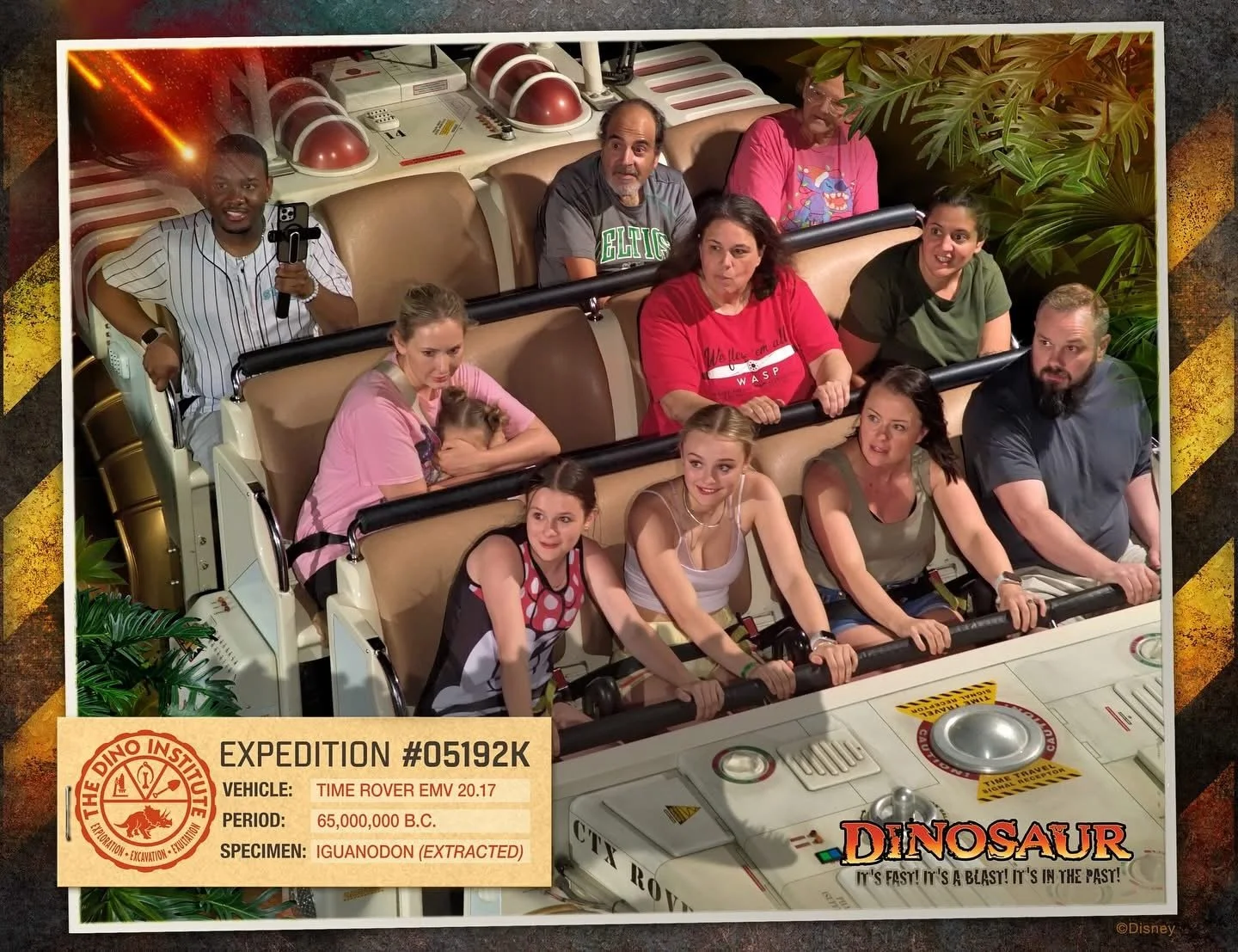 A group of people ride a roller coaster at a theme park. The coaster has a space-themed design, with a control panel and a sign that says 'Dinosaur' and 'It's fast! It's a blast! It's in the past!'
