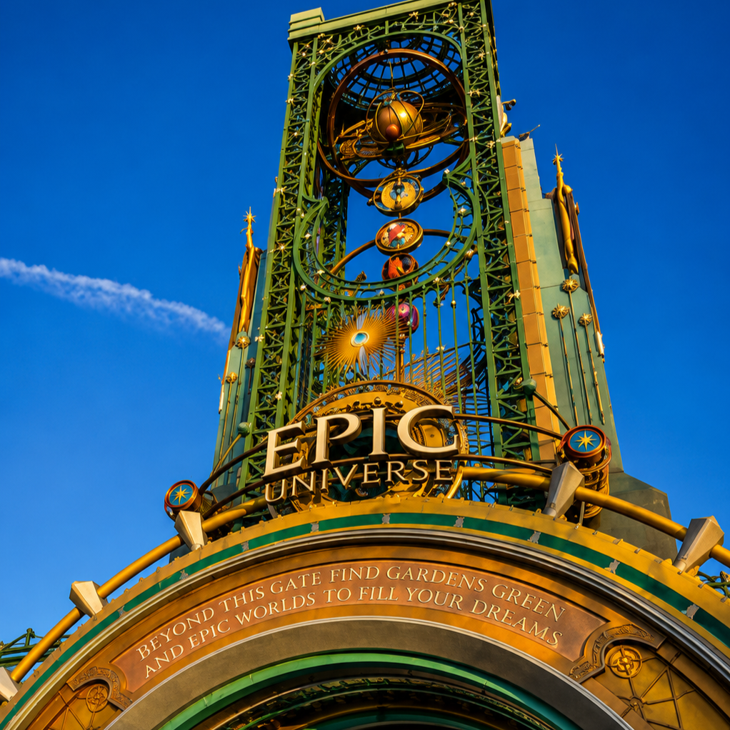 Close-up of the Epically designed entry sign for Epic Universe theme park, featuring green and gold decorative elements and a blue sky background.