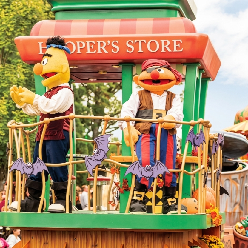 Costumed characters of Bert and Ernie from Sesame Street standing on a parade float decorated for Halloween with bats and pumpkins, labeled 'Hopper's Store'.