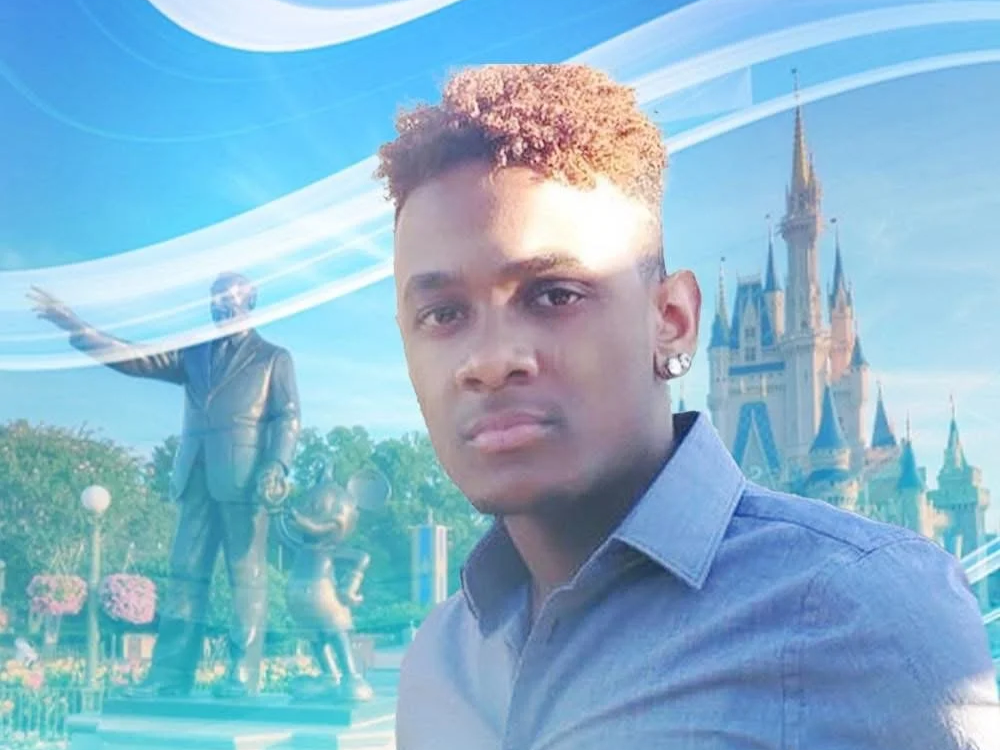 A young man with curly hair taking a selfie in front of a Disney castle and a statue of Walt Disney.