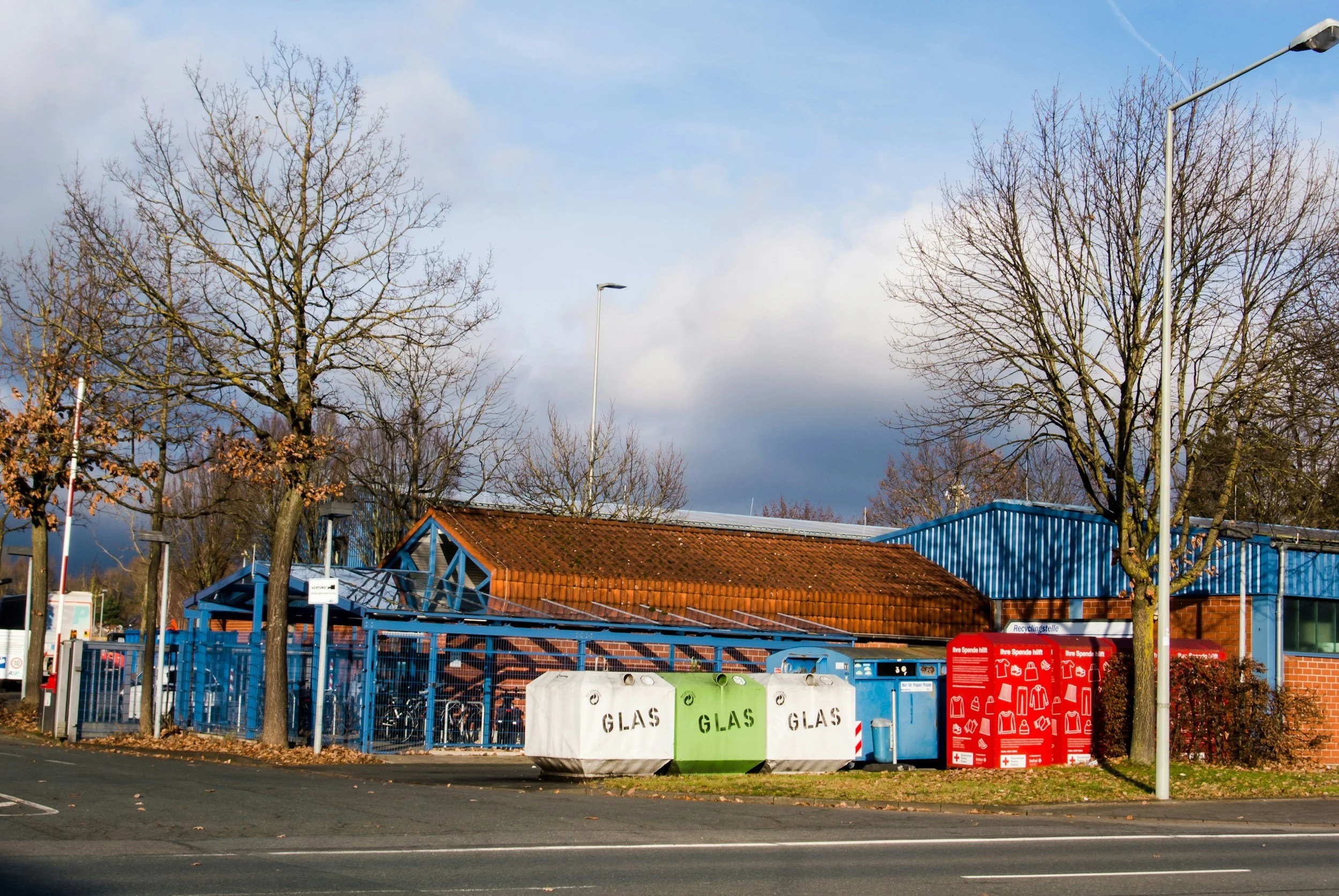 View of a recycling station with containers labeled for glass, surrounded by trees without leaves under a partly cloudy sky.