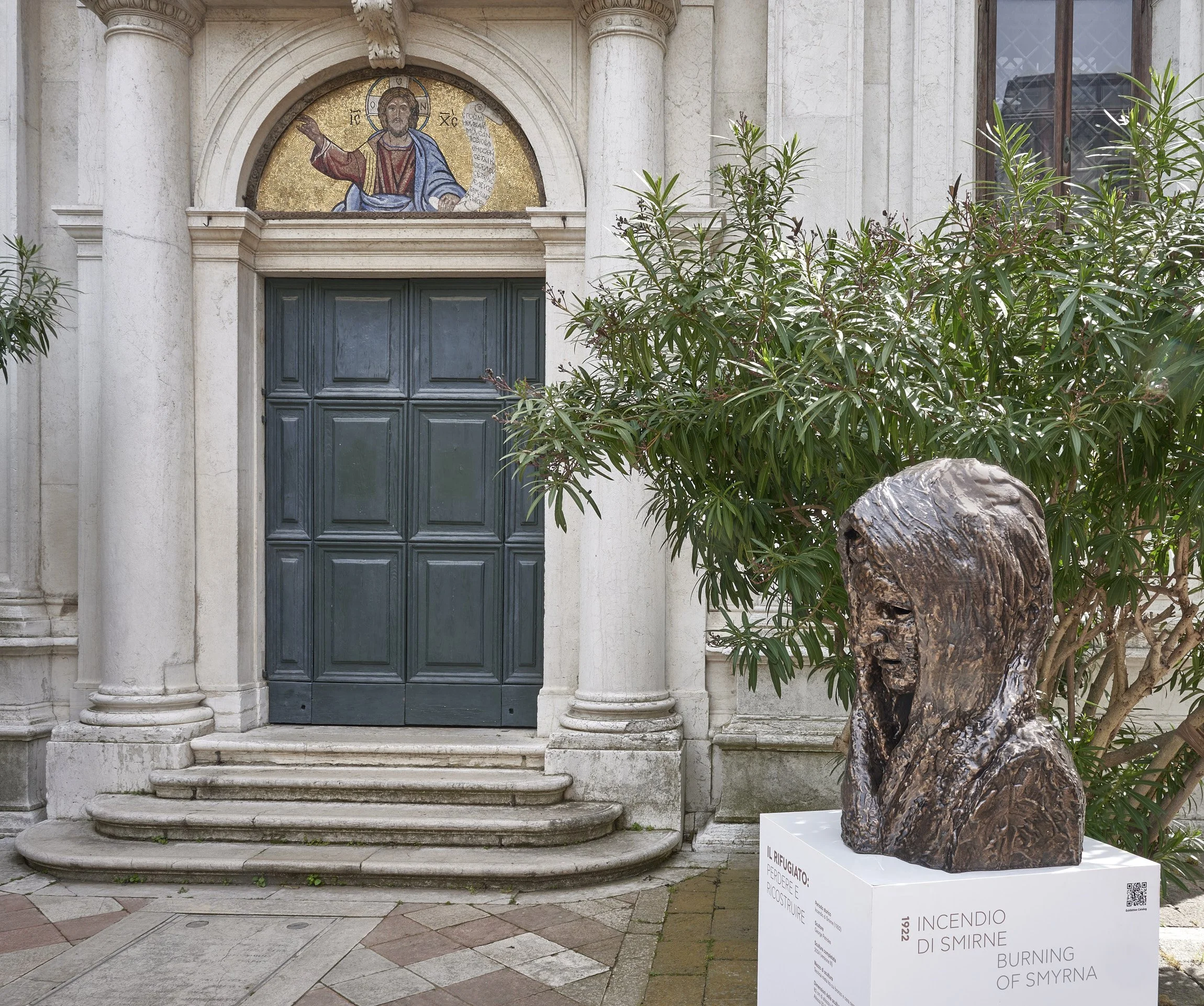 A stone sculpture of a woman with her mouth open, placed on a white pedestal outside a building with a black door, green plants, and a mosaic image of Jesus Christ above the door.