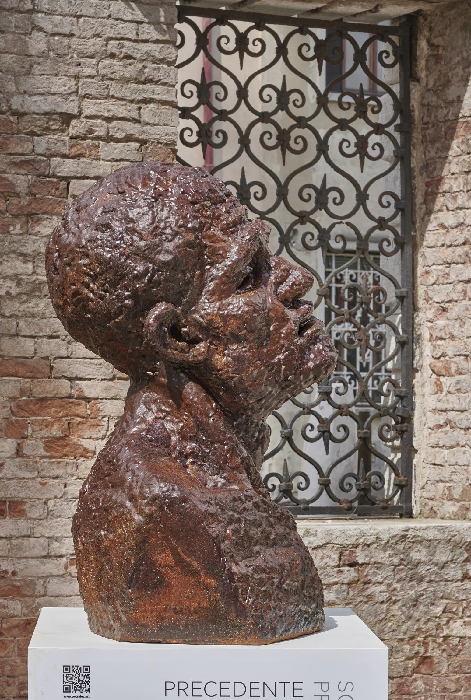 A bronze bust sculpture of a woman with short hair, set against a brick wall and ornate metal window grille background. The sculpture is displayed on a white pedestal with the word 'PRECEDENTE'.
