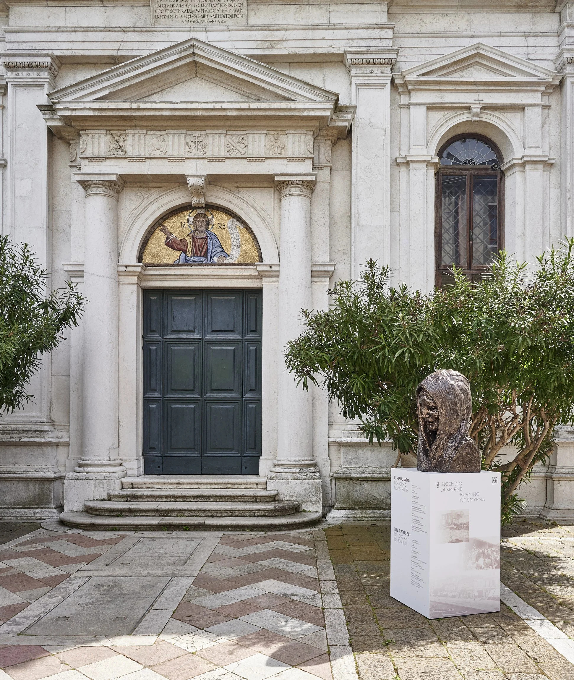 White stone building with a dark green door, a mosaic of Jesus Christ above it, and a plaque in front of a sculpture of a face on a tree next to it.