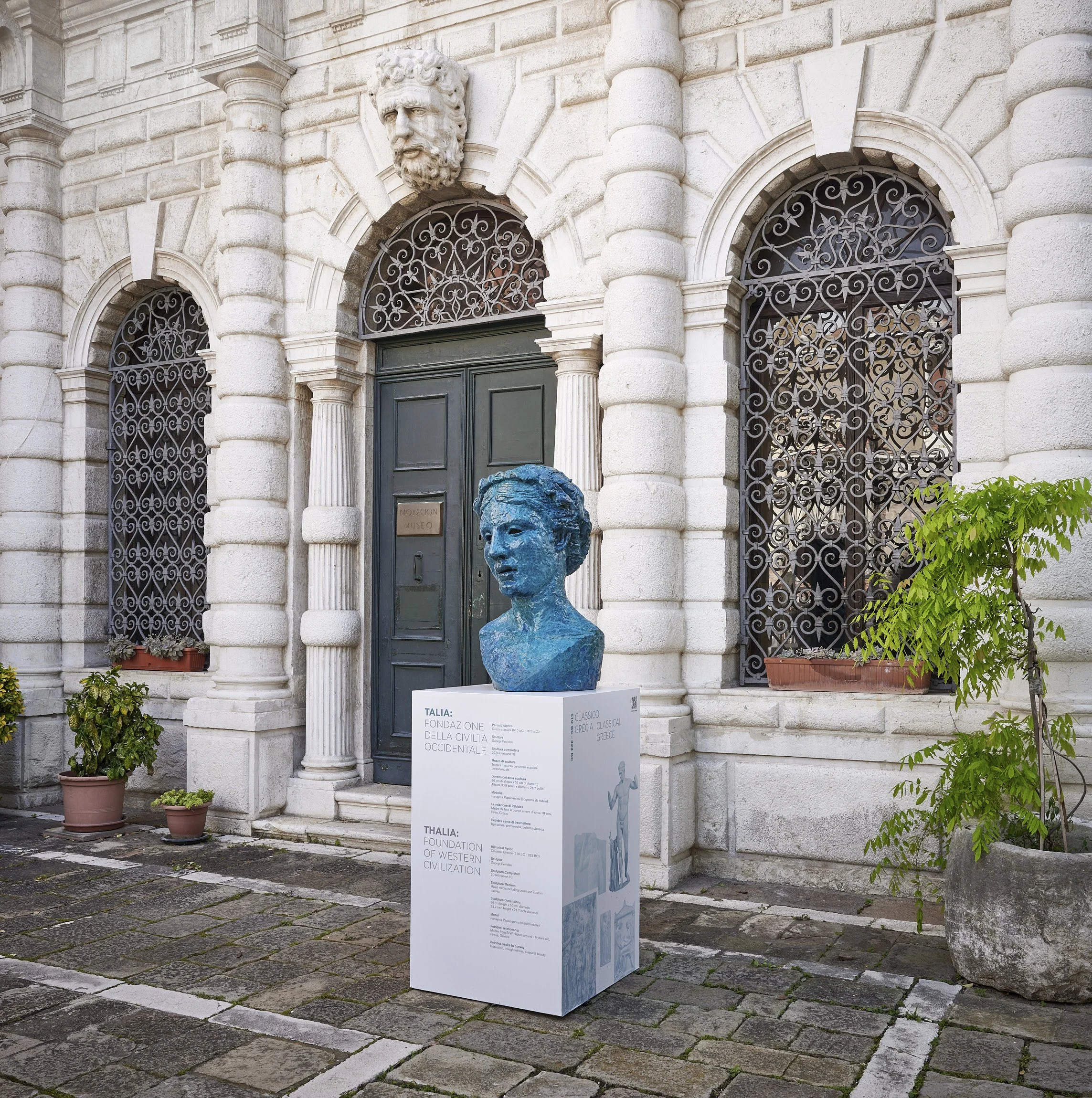 A bust sculpture of a woman painted blue displayed outdoors in front of a historic building with arched windows and decorative iron grilles. There is a white informational sign at the base of the sculpture with text about the foundation of Western ci