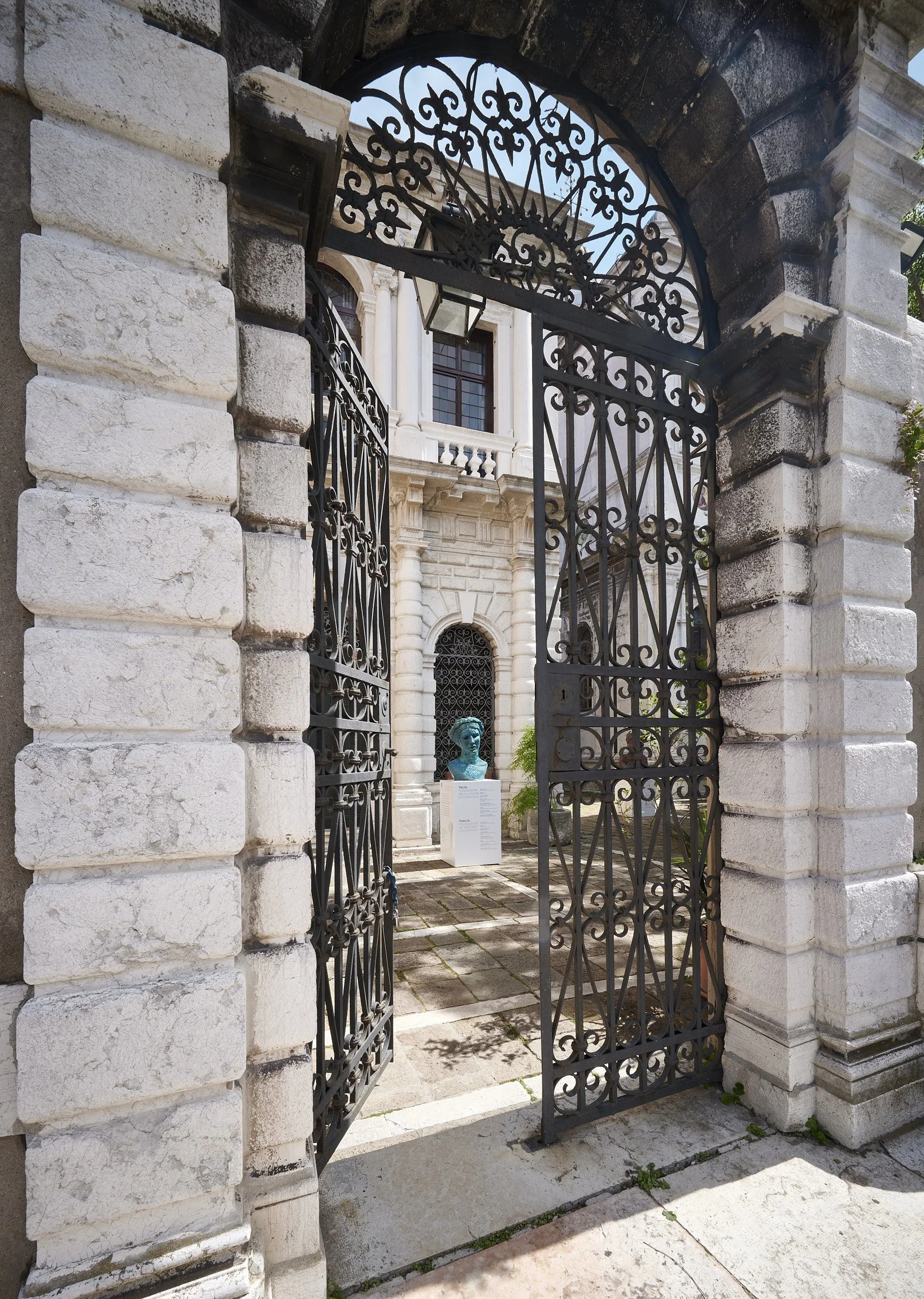 View through a black decorative wrought-iron gate into a historic stone building courtyard with a statue and informational signage.