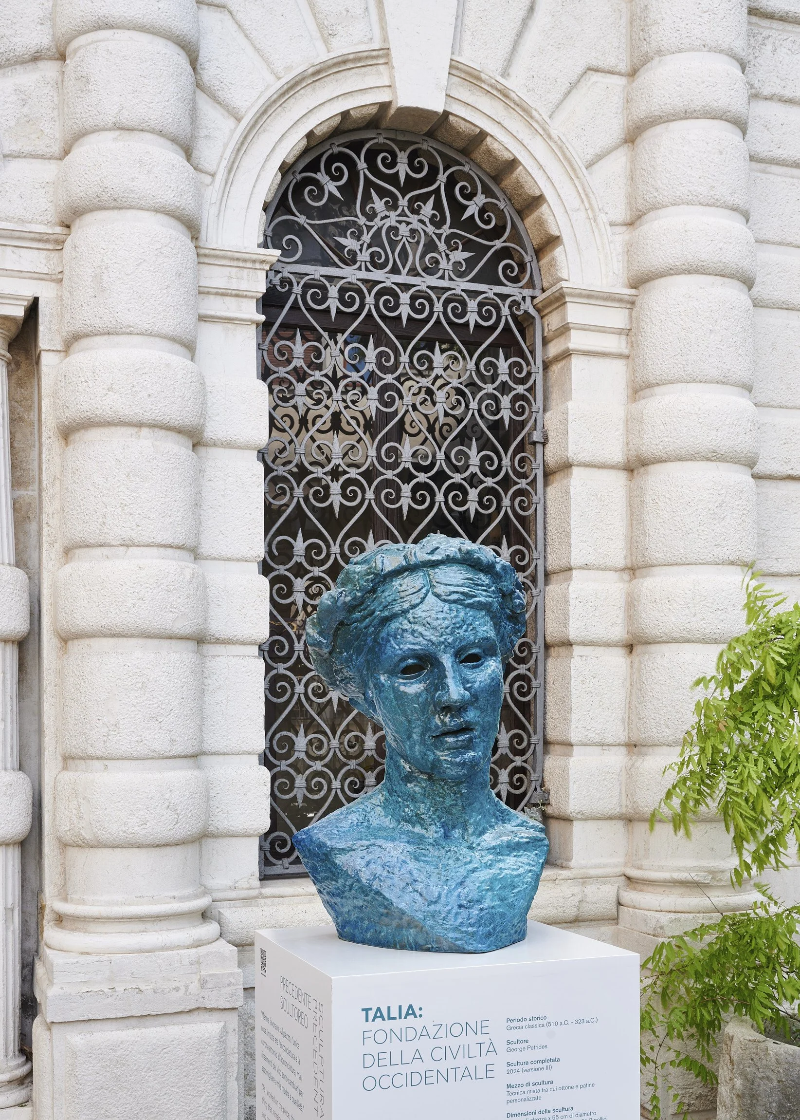 Blue bust sculpture of a woman in front of an ornate iron gate and stone building, with an information plaque below in Italian.