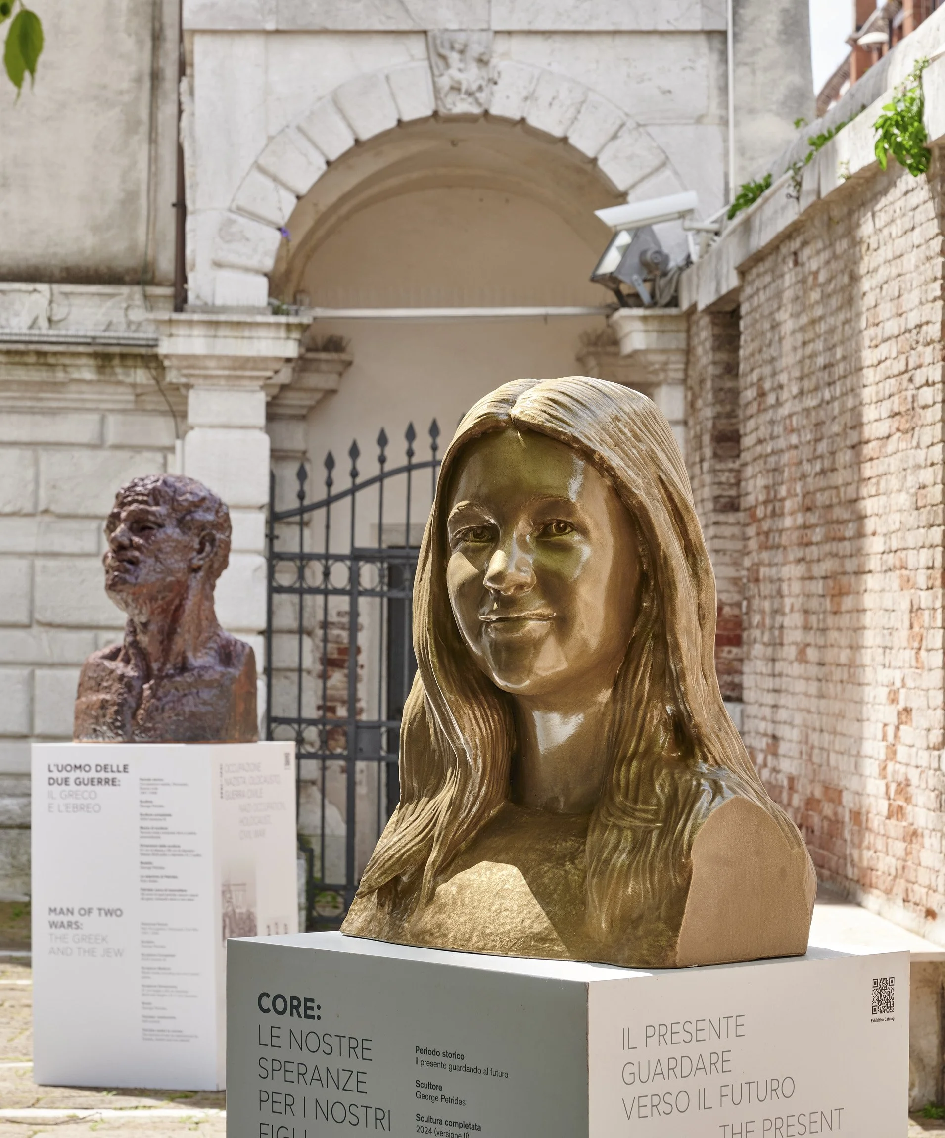 Bronze bust of a young woman with long hair, displayed outdoors with a historical brick wall and archway in the background.