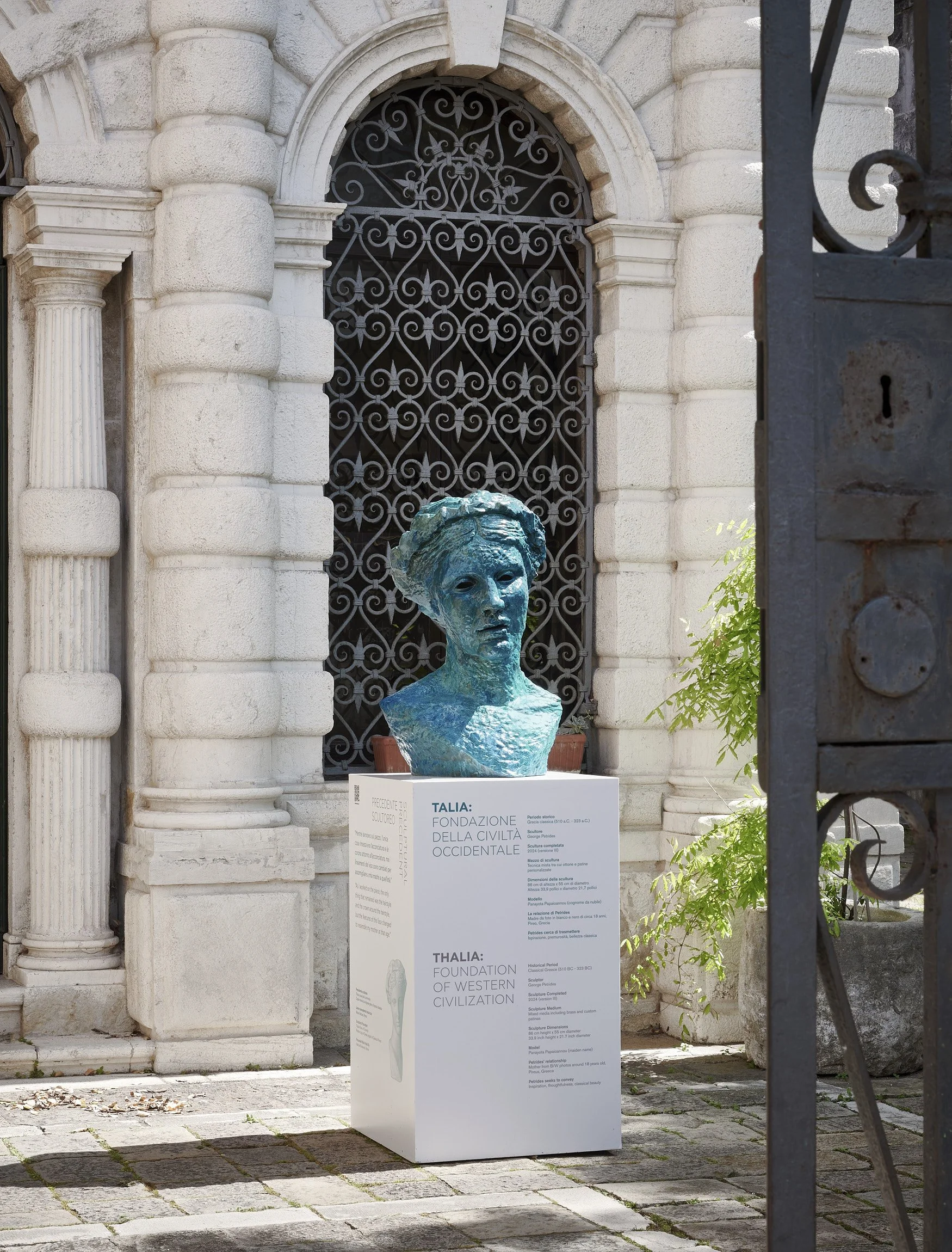 A bust of a woman made of bronze, displayed on a white pedestal, in front of a historic stone building with arched windows and iron grille bars, partially obscured by a black wrought-iron gate.