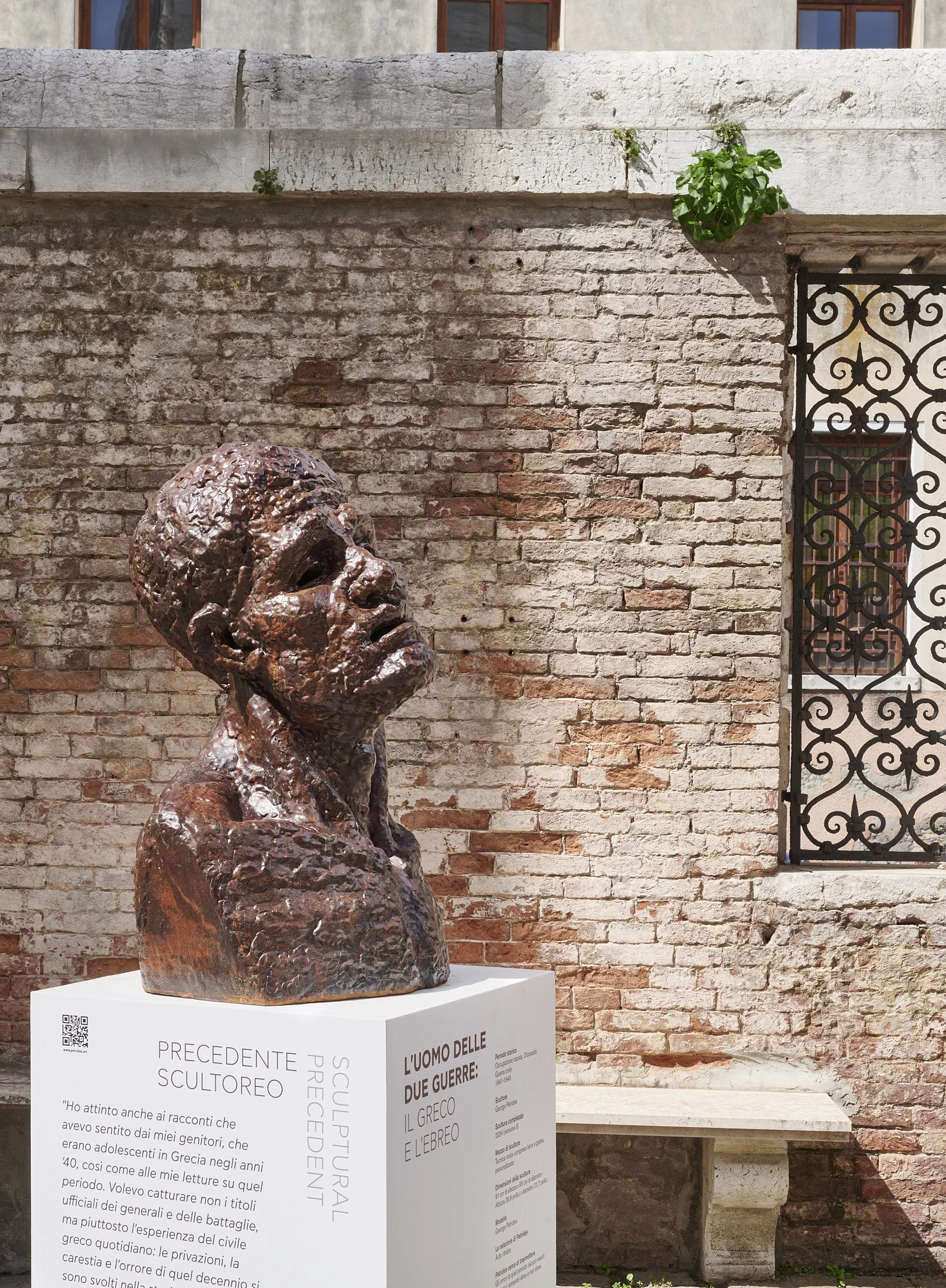Bronze bust sculpture of a man on a white pedestal outside against a brick wall, with a decorative iron window on the right and some green plants on the top left.