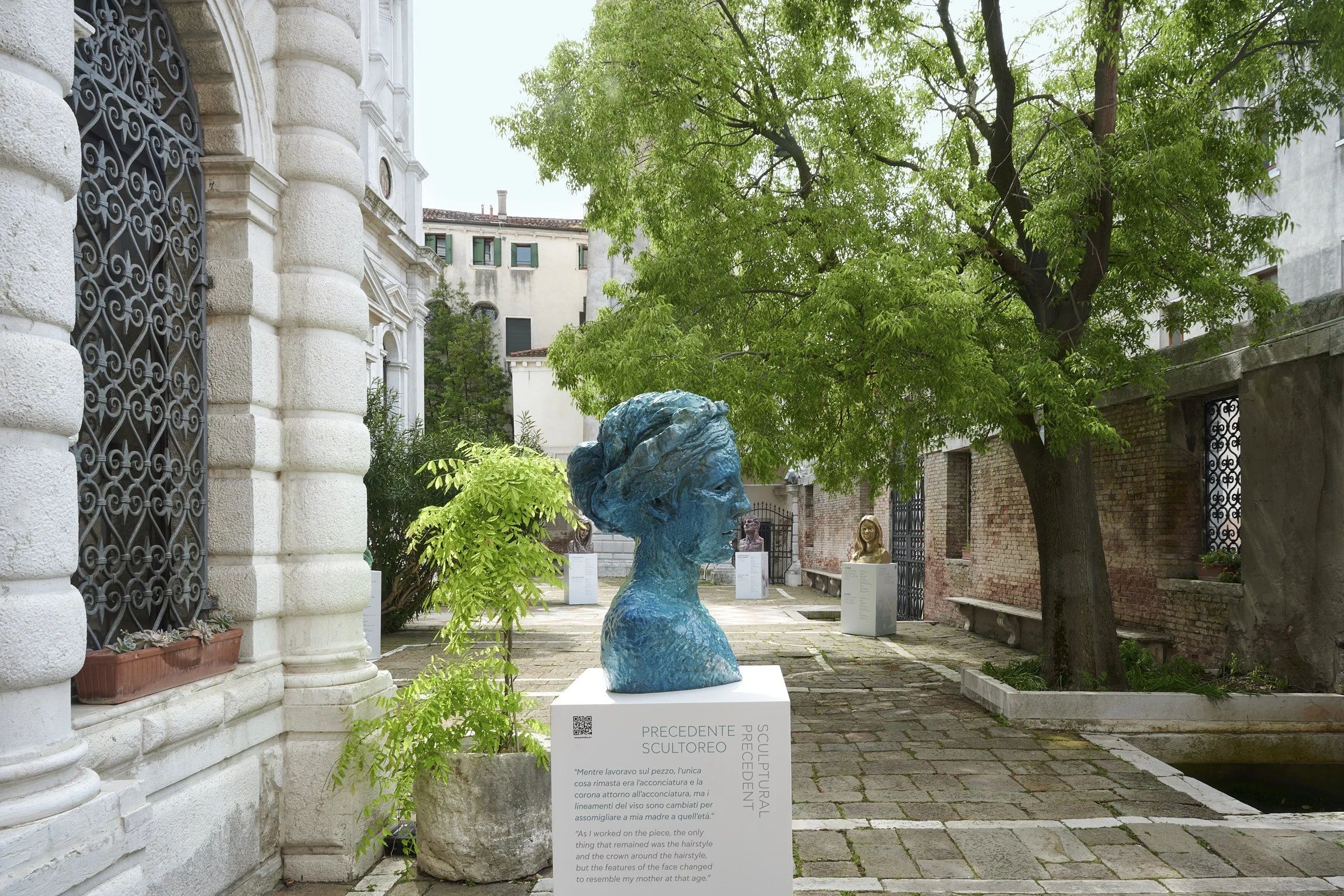 Outdoor sculpture exhibit with a blue bust of a woman in a courtyard, surrounded by trees, greenery, and historic buildings.