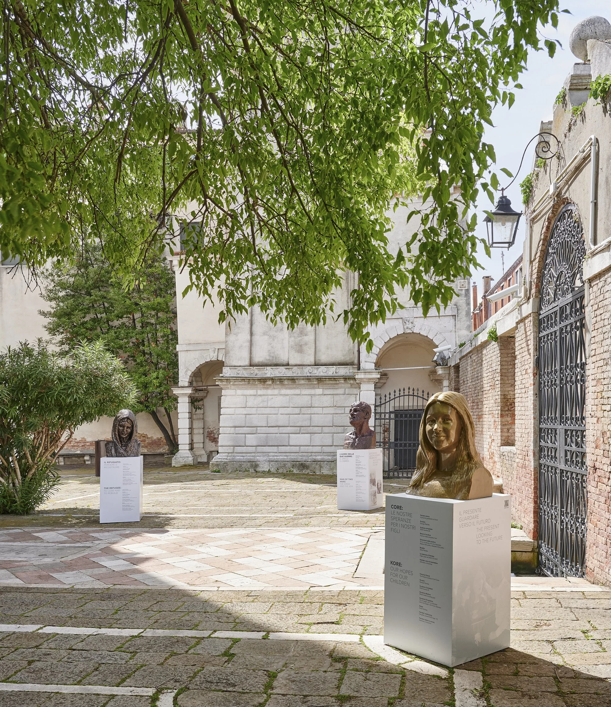 Outdoor art display with three bust sculptures on pedestals, set in a courtyard with cobblestone paving, surrounded by trees and brick walls, with informational plaques on the pedestals.