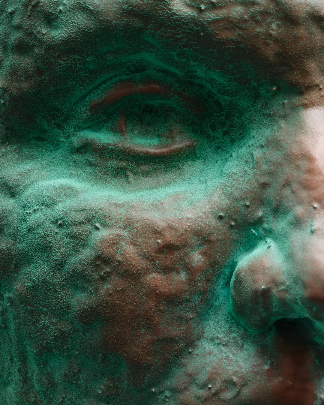 Close-up of a weathered bronze sculpture's face, focusing on the eye and cheek, with a textured greenish patina.