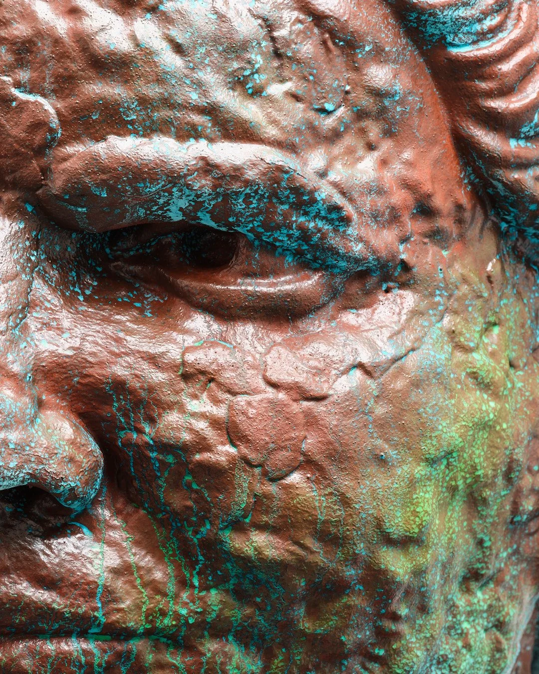 Close-up of a bronze sculpture of a person's face with blue and green paint splatters and streaks.