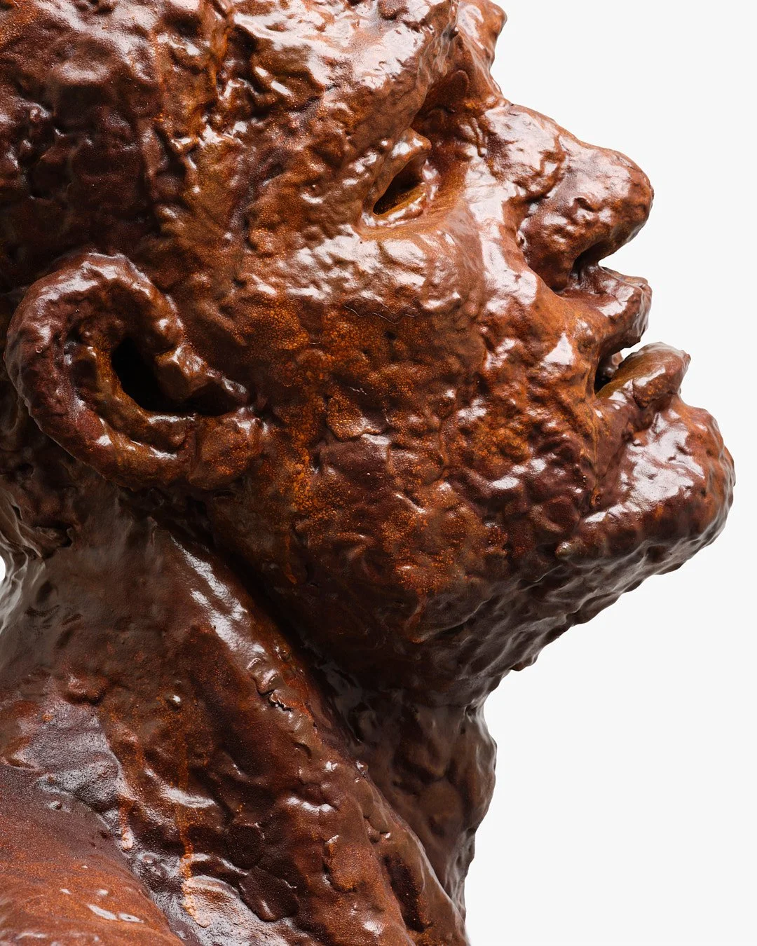 Close-up of a bronze statue of a man's face with textured features, looking upwards, against a plain white background.