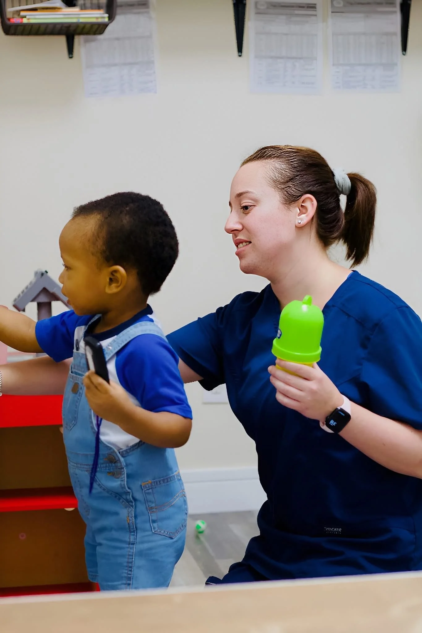 A woman with brown hair tied in a ponytail and wearing a dark blue uniform is kneeling down, holding a green sippy cup, and engaging with a young boy wearing a blue shirt and denim overalls. They appear to be in a classroom or daycare setting.