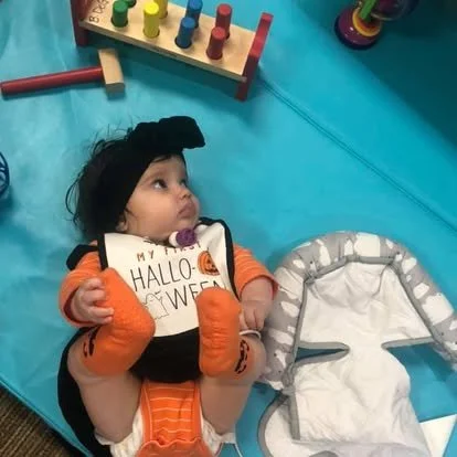 A baby in a pumpkin-themed outfit is lying on a play mat surrounded by toys, including a colorful stacker and a letter matching game.