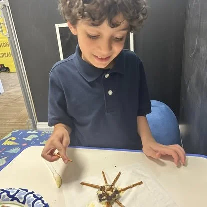 Children's stuffed animal or toy on a table in front of a boy with curly hair, wearing a navy blue polo shirt, sitting at a table with a colorful mat.