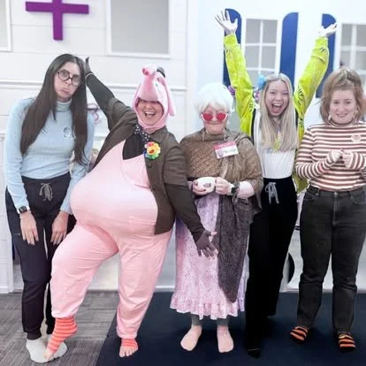 Five women standing and smiling, with two dressed in humorous costumes; one as a pig and the other as an elderly woman, in a brightly decorated room with windows and a cross symbol.