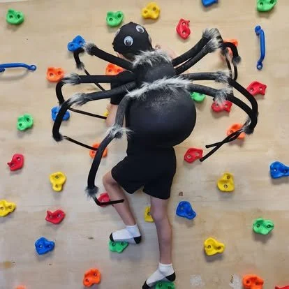 Child in a spider costume climbing a colorful indoor rock wall with climbing holds.