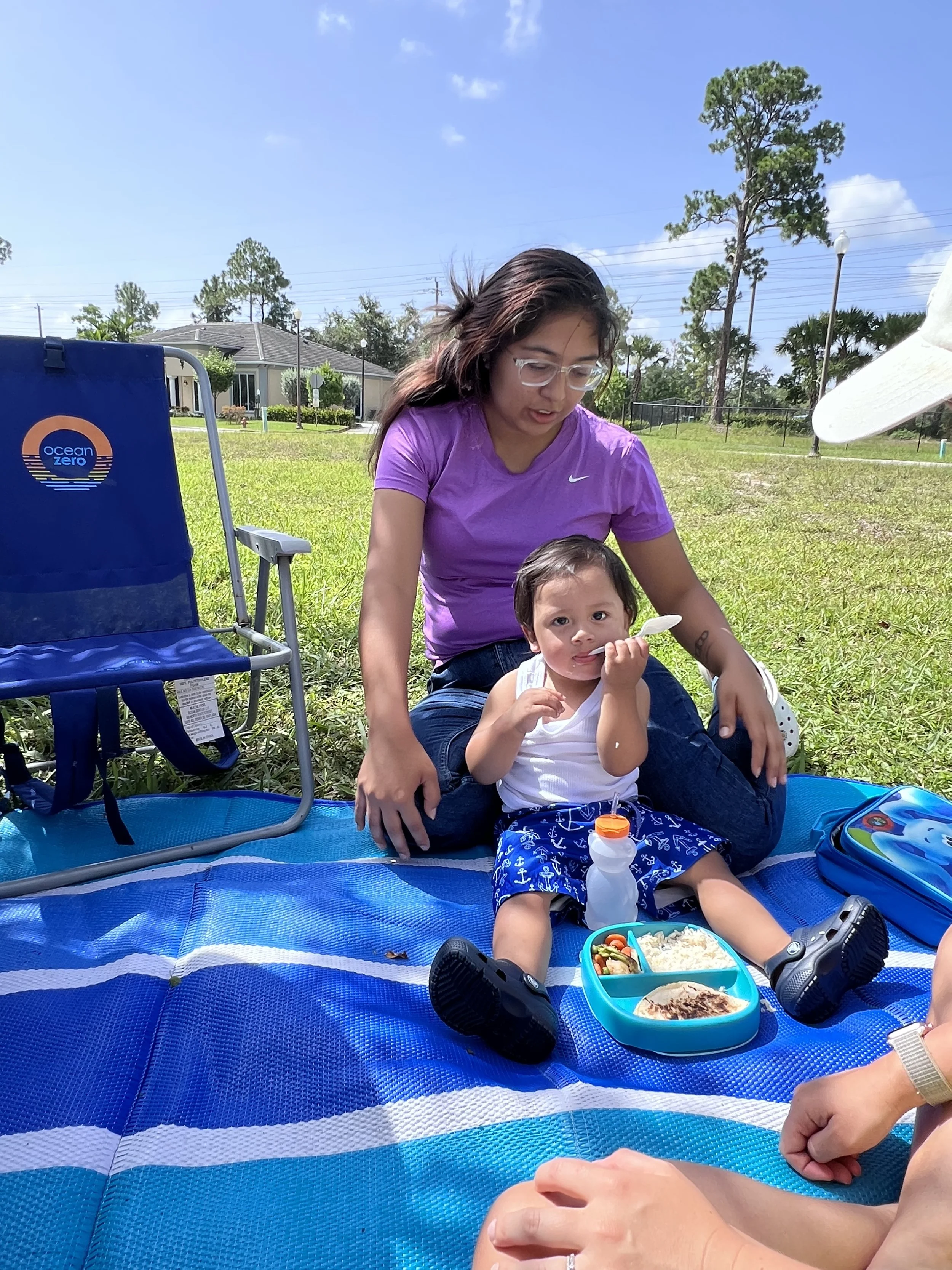 A woman and a young child sitting on a blue picnic blanket outdoors on a sunny day, with the woman helping the child. There is a divided meal container, a water bottle, and a chair nearby. The background features grass, trees, and suburban houses.