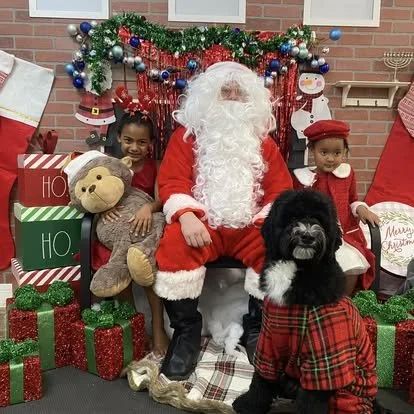 Santa Claus sitting with two children, one holding a teddy bear, in front of Christmas decorations, including gifts, stockings, and holiday figures.