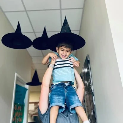 Child dressed as a witch with a pointed hat, sitting on an adult's shoulders in a hallway decorated with additional witch hats hanging from the ceiling.