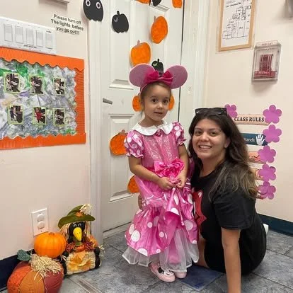 Young girl dressed as Minnie Mouse with pink ears and dress, posing with a smiling woman in a classroom decorated for Halloween.