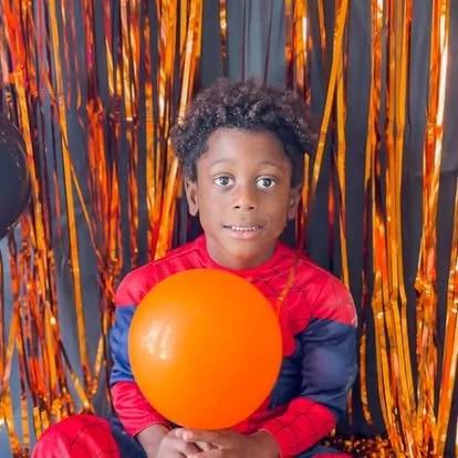 Child sitting in front of a Halloween-themed backdrop holding an orange balloon.