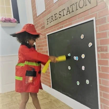 Child dressed as a firefighter drawing on a chalkboard with magnet stickers, in front of a brick wall with a sign that reads 'FIRE STATION'.