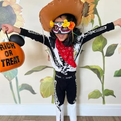Child dressed in a skeleton costume with a sugar skull face paint, wearing a sombrero hat decorated with yellow flowers, holding a pumpkin-shaped bucket labeled "Trick or Treat," and an umbrella, standing in front of a floral wallpaper background.
